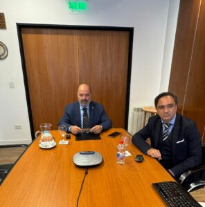 Dos participantes sentados ante una mesa con documentos y un equipo de audio para la reunión.